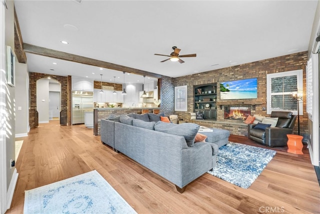 living area with arched walkways, brick wall, ceiling fan, light wood-type flooring, and a fireplace