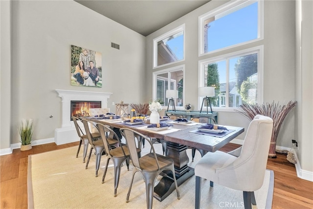 dining room with a towering ceiling, a glass covered fireplace, light wood-style flooring, and baseboards