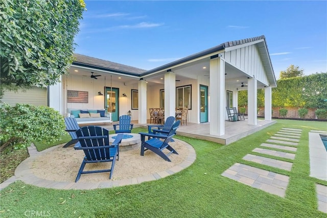 rear view of property with ceiling fan, a patio, a lawn, board and batten siding, and an outdoor living space with a fire pit