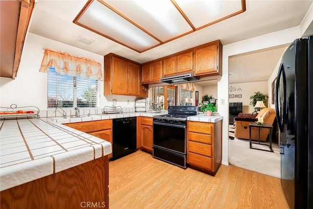 kitchen featuring tile countertops, brown cabinetry, light wood-style floors, under cabinet range hood, and black appliances