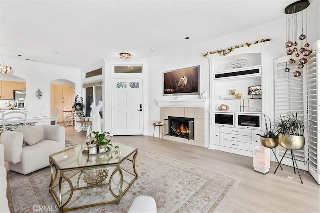 living room with light wood-type flooring, a tiled fireplace, arched walkways, and crown molding