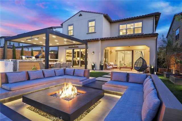 back of house at dusk with stucco siding, a tiled roof, an outdoor kitchen, and an outdoor living space with a fire pit