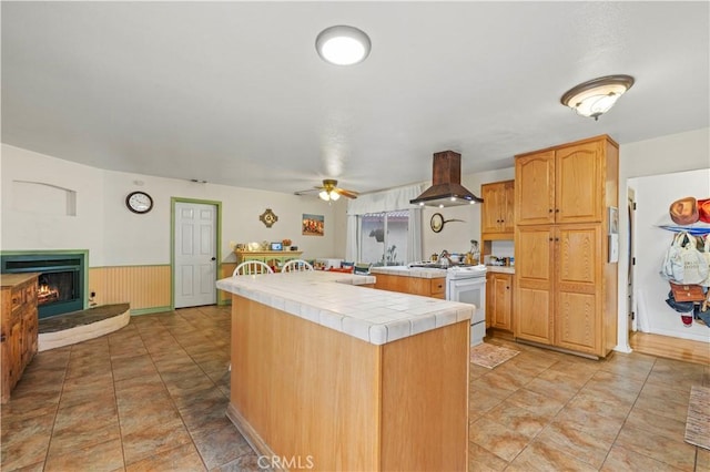 kitchen with a warm lit fireplace, tile counters, wainscoting, island exhaust hood, and white gas stove