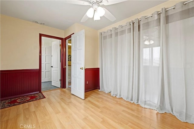 empty room featuring a ceiling fan, a wainscoted wall, visible vents, and wood finished floors