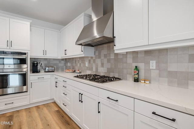 kitchen featuring light wood finished floors, decorative backsplash, appliances with stainless steel finishes, white cabinets, and wall chimney range hood