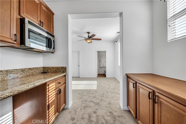 kitchen with light stone counters, light colored carpet, stainless steel microwave, brown cabinetry, and ceiling fan