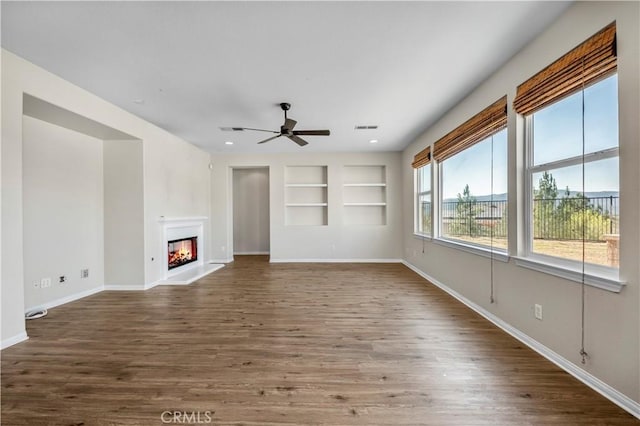 unfurnished living room featuring built in shelves, visible vents, baseboards, dark wood-style floors, and a glass covered fireplace