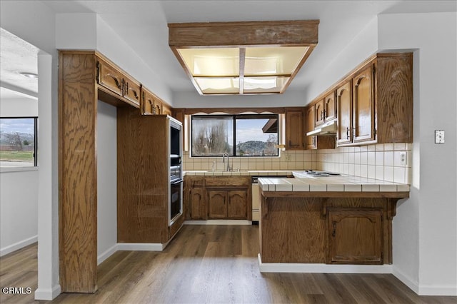 kitchen with tile countertops, dark wood-type flooring, a sink, under cabinet range hood, and backsplash