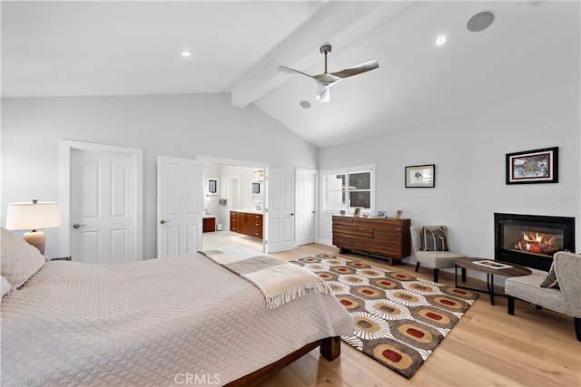 bedroom with light wood finished floors, connected bathroom, a glass covered fireplace, ceiling fan, and beamed ceiling