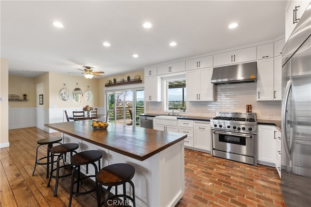 kitchen with white cabinets, wainscoting, butcher block countertops, stainless steel appliances, and under cabinet range hood