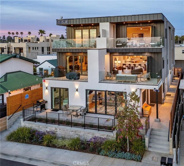 back of property at dusk featuring stucco siding, board and batten siding, a patio area, a balcony, and stairs