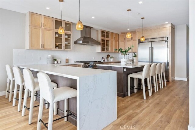 kitchen with a peninsula, stove, decorative backsplash, wall chimney exhaust hood, and stainless steel built in refrigerator