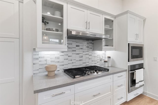 kitchen featuring glass insert cabinets, under cabinet range hood, decorative backsplash, stainless steel appliances, and white cabinetry