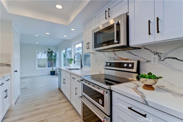 kitchen featuring light wood-style flooring, a sink, appliances with stainless steel finishes, light stone countertops, and a tray ceiling