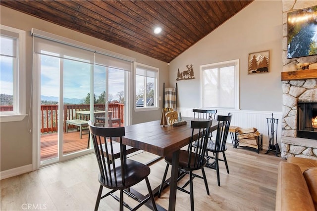 dining area with a stone fireplace, light wood-style flooring, a wainscoted wall, wood ceiling, and vaulted ceiling