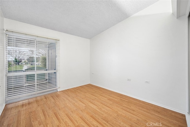 empty room with a textured ceiling, vaulted ceiling, baseboards, and light wood-style floors