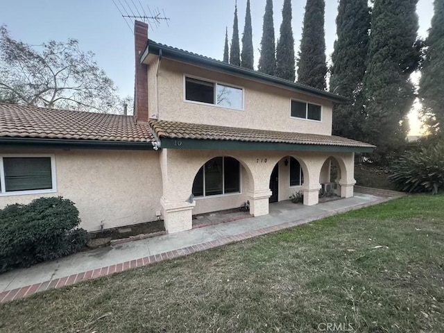 rear view of property with a tiled roof, a lawn, a chimney, and stucco siding