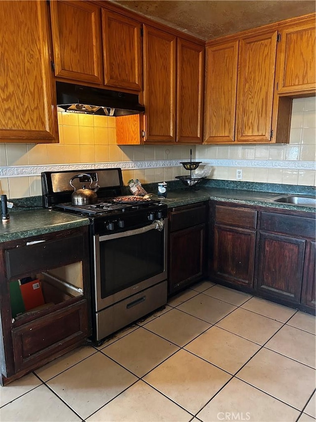 kitchen featuring stainless steel gas stove, dark countertops, a sink, under cabinet range hood, and backsplash