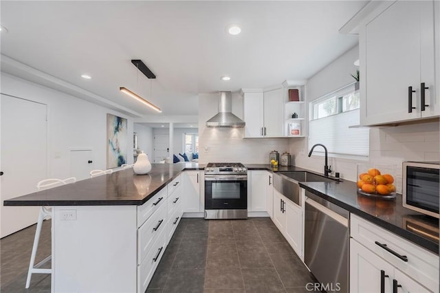 kitchen with stainless steel appliances, dark countertops, a sink, a peninsula, and wall chimney exhaust hood