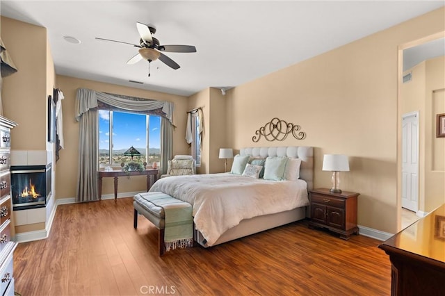 bedroom featuring a ceiling fan, baseboards, a tiled fireplace, and wood finished floors