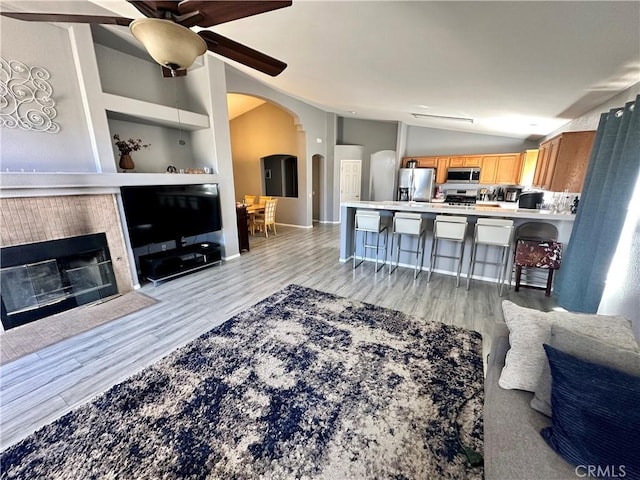 living area featuring arched walkways, a fireplace, a ceiling fan, light wood-style floors, and vaulted ceiling