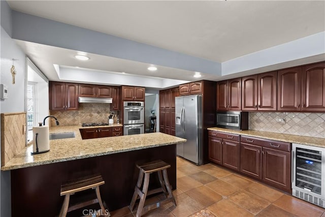 kitchen with wine cooler, a tray ceiling, appliances with stainless steel finishes, a sink, and under cabinet range hood