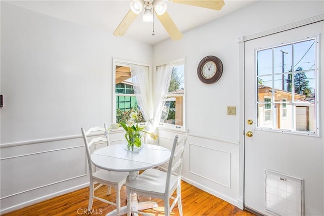 dining room featuring ceiling fan, light wood-style flooring, wainscoting, and a decorative wall