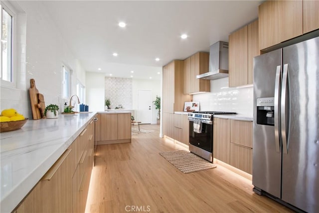 kitchen with light brown cabinetry, appliances with stainless steel finishes, light wood-style floors, wall chimney range hood, and modern cabinets