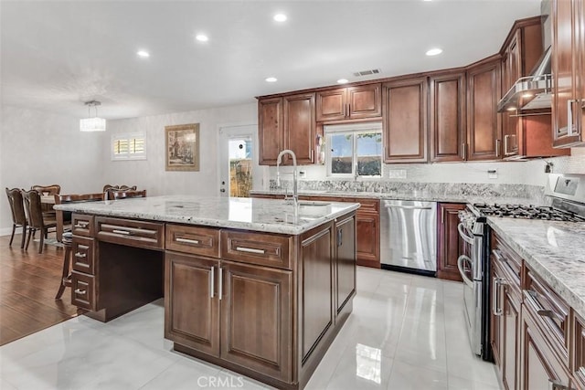 kitchen featuring a wealth of natural light, appliances with stainless steel finishes, a sink, an island with sink, and light stone countertops