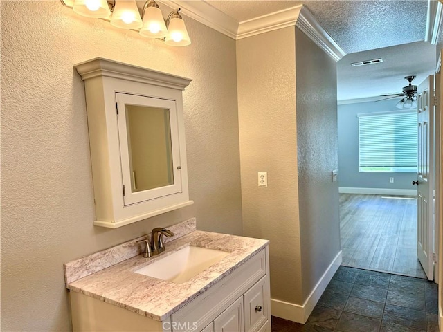 bathroom with ceiling fan with notable chandelier, vanity, visible vents, baseboards, and ornamental molding