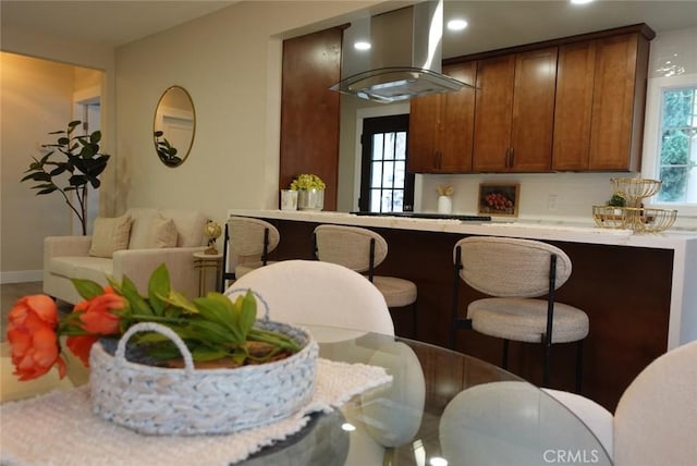 kitchen featuring light countertops, wall chimney range hood, tasteful backsplash, brown cabinetry, and a kitchen bar