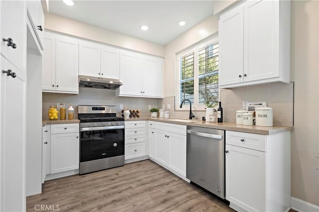 kitchen with light wood finished floors, appliances with stainless steel finishes, under cabinet range hood, white cabinetry, and a sink