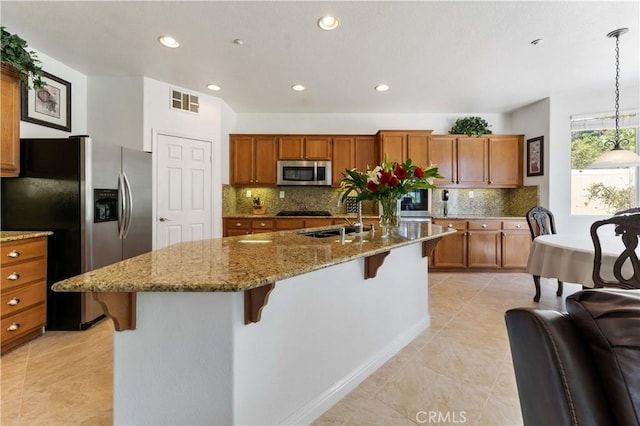 kitchen featuring brown cabinetry, an island with sink, appliances with stainless steel finishes, a kitchen breakfast bar, and light stone countertops