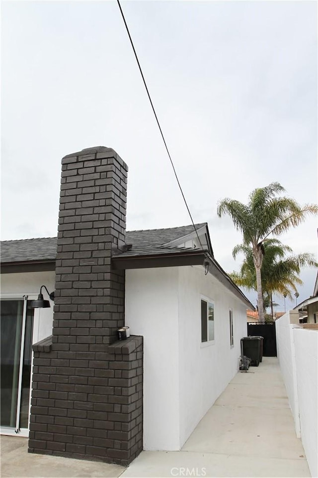 view of side of home with a shingled roof, a patio, a chimney, fence, and stucco siding