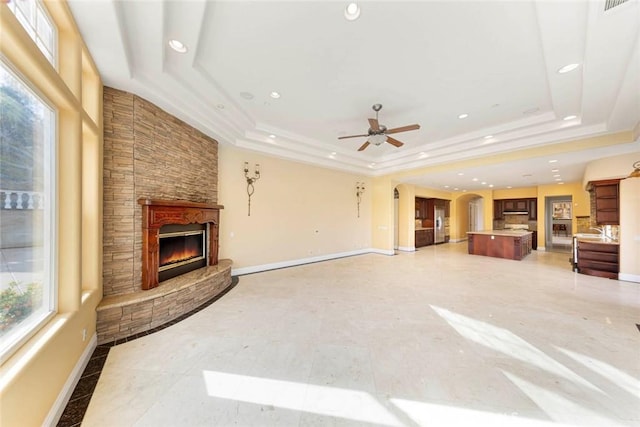 unfurnished living room with arched walkways, a tray ceiling, a stone fireplace, plenty of natural light, and baseboards