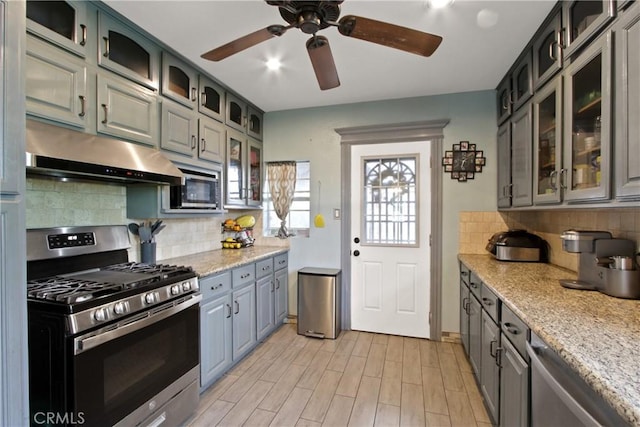 kitchen with light wood-style flooring, glass insert cabinets, appliances with stainless steel finishes, under cabinet range hood, and backsplash