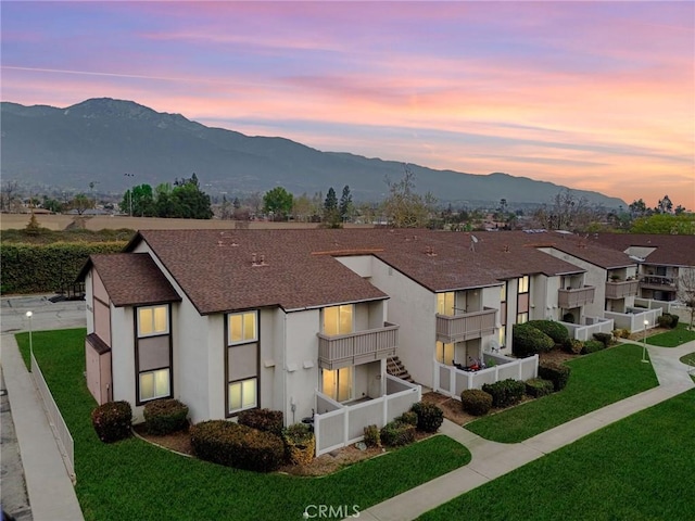 view of property featuring a balcony, stucco siding, roof with shingles, a mountain view, and a front yard