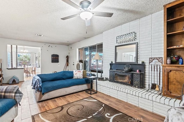 living area featuring visible vents, a glass covered fireplace, a textured ceiling, wood finished floors, and ceiling fan with notable chandelier