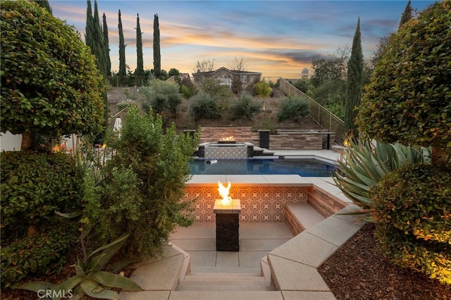 pool at dusk with an outdoor fire pit, a pool with connected hot tub, a fenced backyard, and a patio