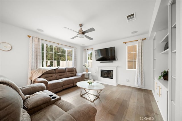 living room featuring light wood-style flooring, visible vents, a ceiling fan, and a glass covered fireplace