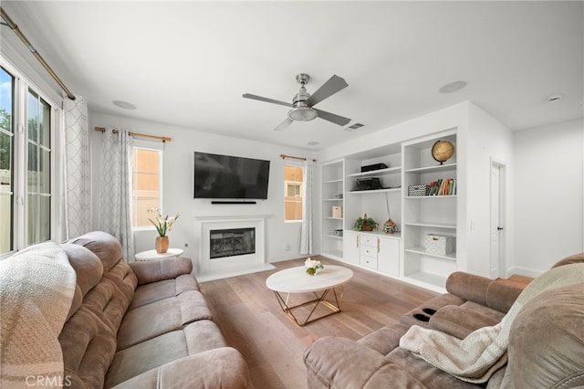 living area with built in features, a fireplace with flush hearth, a ceiling fan, wood finished floors, and baseboards