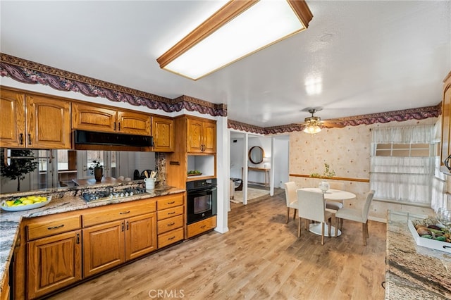 kitchen with under cabinet range hood, black oven, brown cabinets, wallpapered walls, and stainless steel gas stovetop