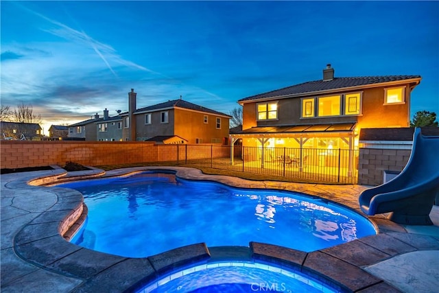 pool at dusk with a pool with connected hot tub, a patio, a water slide, and a fenced backyard