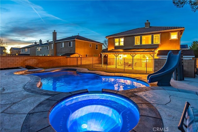 pool at dusk with a patio, a water slide, a fenced backyard, and a pool with connected hot tub