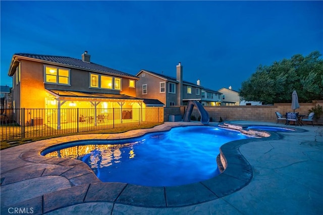 pool at twilight featuring a patio area, a fenced backyard, a pool with connected hot tub, and a water slide