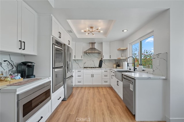 kitchen with appliances with stainless steel finishes, backsplash, a tray ceiling, wall chimney range hood, and a sink