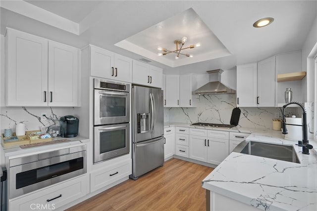 kitchen with a sink, appliances with stainless steel finishes, light stone countertops, wall chimney exhaust hood, and a tray ceiling