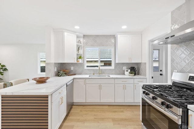 kitchen with light wood finished floors, stainless steel appliances, a peninsula, a sink, and wall chimney range hood