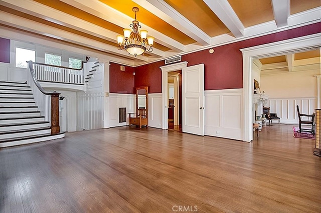 unfurnished living room featuring a wainscoted wall, beamed ceiling, wood finished floors, an inviting chandelier, and stairs