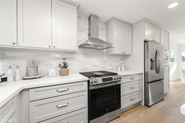 kitchen featuring light wood-style flooring, light countertops, appliances with stainless steel finishes, wall chimney range hood, and tasteful backsplash